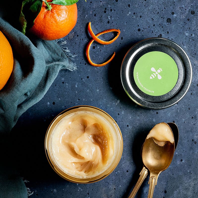 Pot de miel bio de ronce Maison Fedon ouvert, avec cuillère dorée et zestes d’orange.