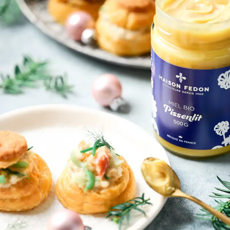 Un pot de miel bio de pissenlit Maison Fedon avec des choux apéritif sur des assiettes et une cuillère de miel.