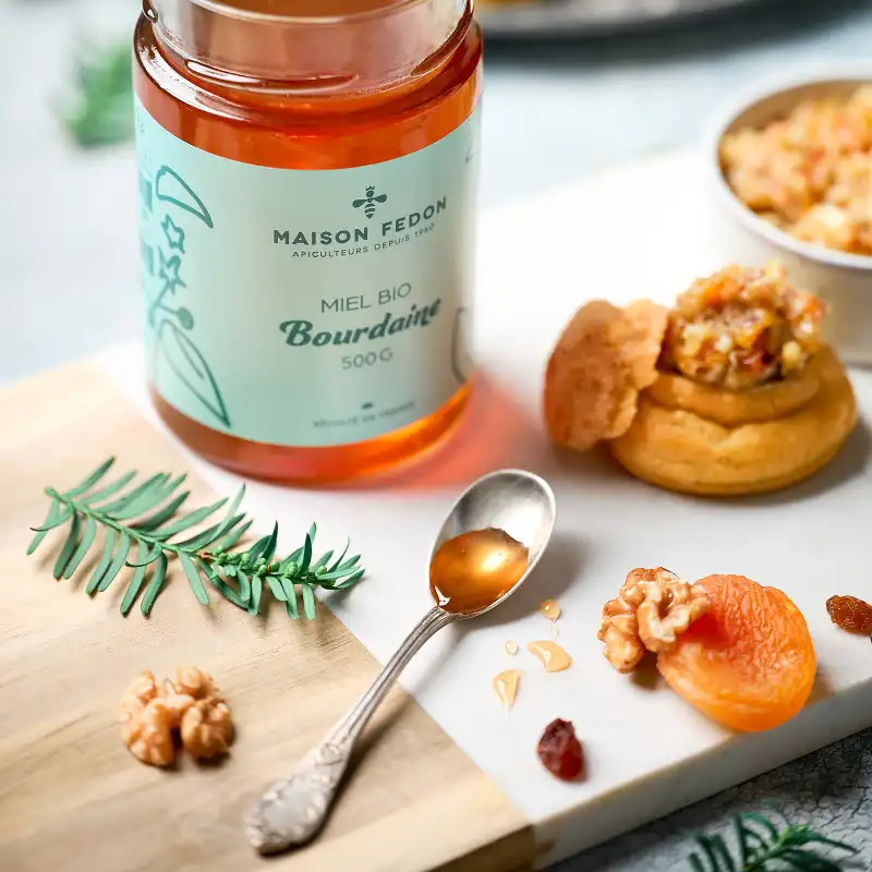 Un pot de miel bio de bourdaine Maison Fedon avec un choux apéritif garni et une cuillère de miel sur une planche.
