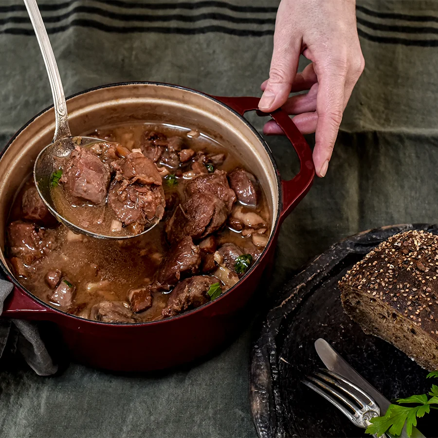 Joue de porc à la bière et au miel dans une cocotte rouge, servie à la louche sur nappe vert foncé.