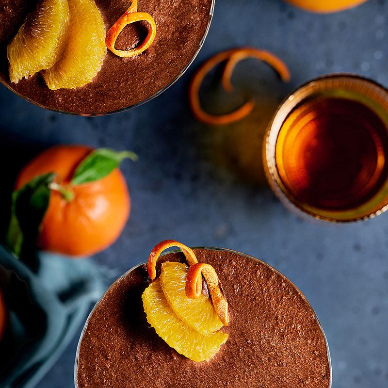 Gros plan sur deux coupes de mousse au chocolat noir, décorée de zestes et quartiers d’orange, et verre de sirop.
