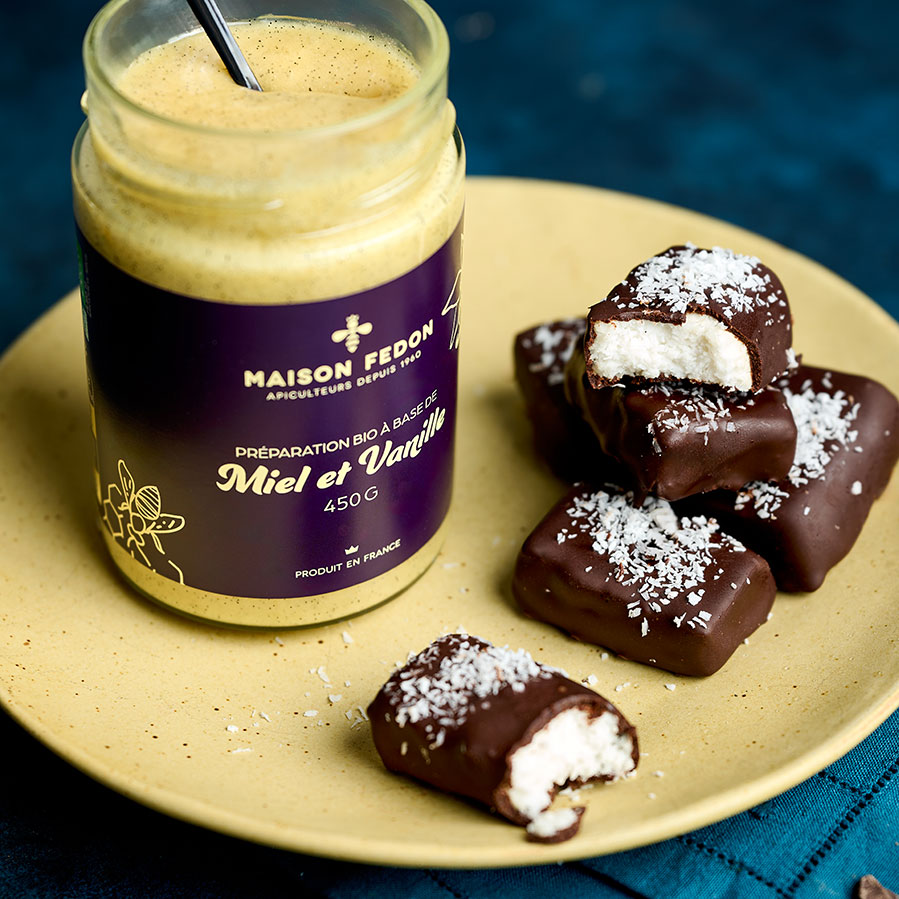 Bounty maison au chocolat noir et à la noix de coco, dont un coupé en deux révèle son cœur fondant, à côté du pot Miel & Vanille Maison Fedon.